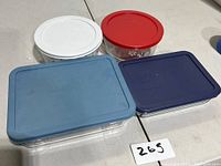 Four Pyrex glass storage bowls with lids in white, red, blue, and navy blue colors arranged on a floor.