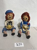 Full view of both Raggedy Ann and Andy figurines seated side by side on a tiled floor against a white wall.