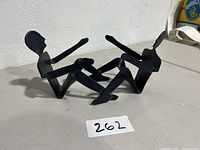Front and side perspective showing black metal abstract sculpture of a couple with bent knees and extended arms.