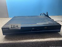 Front angled view of Sony Blu-ray DVD player showing brand, Blu-ray logo, and front panel with buttons and disc tray. Device is black with visible fingerprints and dust.