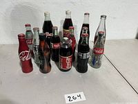 Front view displaying a collection of 14 glass Coca Cola bottles with different sizes, shapes and labels.