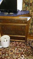 Dresser front with four drawers, medium brown wood, round drawer knobs, placed on patterned carpet with a lamp in front and a TV screen on top.
