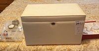 White jewelry box closed on patterned fabric surface with plastic container holding small colorful jewelry-making items, silver pocket watch and chain, necklace on a red holder tray.