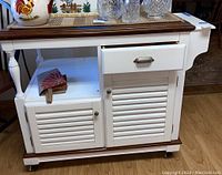 Front view of Coaster kitchen buffet cart showing two louvered cabinet doors, one drawer with silver handle, white body, dark wood top and base, and caster wheels.