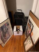 Framed artworks and two wheeled suitcases on floor with box of ceramic collectibles