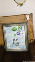 Framed painting of a hummingbird flying toward flowers, with a silver frame and slightly domed glass.