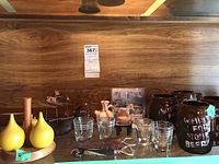 Photo showing yellow ceramic condiment containers on a wooden tray, clear short glasses, brown ceramic jars with text, and various small cups and corkscrews on a wood shelf