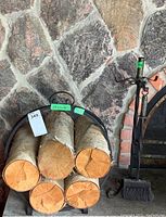 Wrought iron brush and two tools (poker and tongs) placed upright next to a stone fireplace with some firewood logs stacked nearby. Brush has a wooden handle and metal bristles.