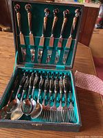 Open black case showing six knives in lid and six forks and six spoons in lower tray against green velvet lining