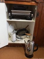 Photos of kitchen appliances and cookware inside a wooden cabinet, showing Cuisinart panini grill, Hamilton Beach coffee percolator, glass measuring cup, metal mixing bowls, fry pans, grinder, and plates.