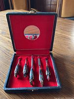 Five-piece manicure set in original box with red velvet lining and mirror in lid, showing ornamental handles shaped like hands and birds with intricate enamel decoration in black, red, white and turquoise