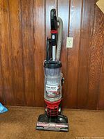 Full view of the MaxLife upright vacuum cleaner standing against a wood-paneled wall showing the vacuum's hose, handle, and base unit.