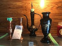 Four decorative metalware items arranged on a wooden surface: two copper bud vases, an Arabic silver inlay coffee pot, and a black vase with silver floral inlay. Some wear and small chips visible on bases.