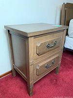View of wood nightstand showing two drawers with metal handles, wood grain finish, and tapered legs with decorative accents.