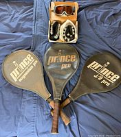 Three Prince tennis racquets in their cases, ankle weights in white and black, and a ski mask on a blue fabric surface.