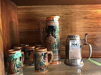 Photo showing six ceramic steins, one tall ceramic jug, and one pewter lidded stein grouped on a wooden shelf.