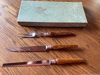 Set of three carving tools with wooden handles and steel blades lying flat with original box above them. Box is greenish and worn with stains and marks.