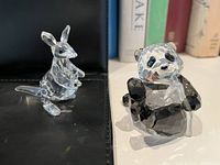 Clear Swarovski crystal kangaroo and black-and-clear crystal panda figurines side by side on a black surface with books in background.