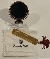 Compact mirror standing on a clear stand, gold-tone frame with Finesse Le Model branding, displayed on top of branded fabric pouch along with gold-tone comb with red tassel.