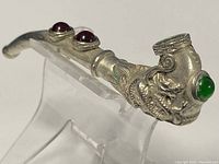 Side angle of a curved vintage Chinese Tibetan silver pipe with dragon design and red and green stones on a clear stand.