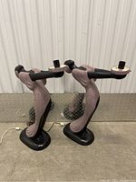 Pair of stylized female figurine table lamps photographed standing side by side on a concrete floor with a metal backdrop.