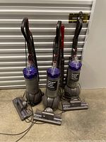 Three upright Dyson Ball vacuums with grey bodies, purple cyclone tops, clear dust bins, and ball-shaped bases on storage floor.