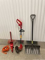 Photo of the three garden tools lined up against a white wall: electric hedge trimmer, electric string trimmer, and snow shovel.