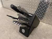 Knife set in black block on concrete floor against metal backdrop. Contains six knives and the block; some slots empty.
