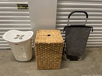 Full view of three laundry items side by side: white plastic basket with lid, woven wicker basket, and grey fabric wheeled cart.