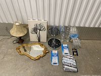 Table lamp, floral wall art, gold mirror, black clock, glassware, packaged bulbs and hardware, and striped textile item on the floor