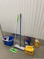 View of complete lot showing cleaning supplies: 2 mops, bucket with wringer, and 3 detergent bottles on concrete floor against white corrugated wall.