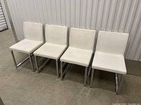 Four white upholstered dining chairs with chrome cantilever bases aligned side by side against a wall, showing front and side views.