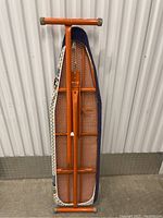 Photo of vintage folding ironing board with orange metal frame and padded patterned cover, standing upright against a corrugated wall.