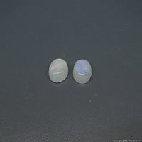 Photo of two oval cabochon rainbow moonstones on a dark background, showing the white base and blue flashing characteristic of the stone.