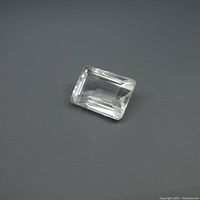 Clear white emerald cut quartz gemstone placed on a gray background showing the facet angles and clarity.