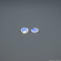 Photo showing two oval blue moonstones resting on a gray surface, displaying a blue sheen.
