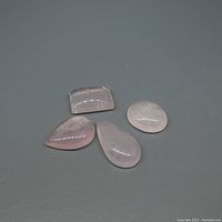 Four rose quartz cabochon cut gemstones on a grey surface showing their shape and color: oval, pear, square, and round/oval shapes.
