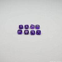 Photo showing eight square cushion amethyst gemstones laid out on a grey background to display their color and cut.