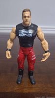 Front view of WWE Chris Benoit action figure showing black sleeveless shirt with 'Toothless Aggression', red pants, and black boots.