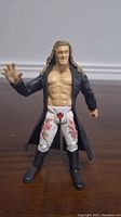 Front view of WWE Edge action figure showing torso, face, and front of attire including coat, tights with red flame designs and cross symbol, and black boots.