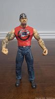 Front view of WWE Undertaker action figure showing red sleeveless shirt with graphic, tattooed arms, blue jeans, black boots, and bandana.