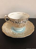 Front and angled view of white china tea cup with gold floral detailing, scalloped edge and gold-trimmed handle on matching saucer.