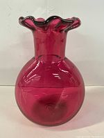 Full view of the cranberry glass vase showing its bulbous body and ruffled top edge, highlighting the deep red color and glossy finish.