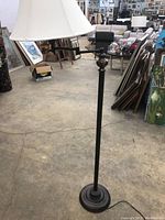Full view of floor lamp standing on concrete floor in a warehouse or store setting, with framed pictures and other items in the background.