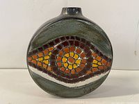 Front view of the ceramic vase showing the oval flattened shape, mosaic tile design in orange, yellow and brown, and the signature in the lower right corner. The background glaze is muted greenish with a small circular neck opening.