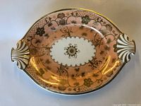 Front view of a vintage decorative dish showing pink porcelain with gold floral patterns and dual shell-shaped gold-accented handles