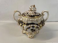 Front view of ornate vintage porcelain sugar bowl with lid and dual gold-handled sides.