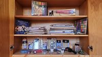 Three shelves filled with various arts and crafts supplies including watercolor pencils, markers, stamps, adhesives, and ink pads.