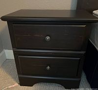 Front view of one black wooden bedside table showing two drawers with round metal knobs, scratches visible on drawer fronts.