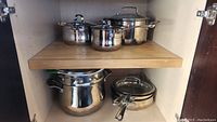 View of seven stainless steel Cuisinart pots and pans including stock pots, saucepans, and a steamer insert inside a kitchen cabinet shelf.